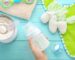 A hand holds a baby bottle filled with milk above a turquoise wooden surface. Nearby are a can of powdered formula with a scoop, a green bib, a pacifier, a towel, and a crocheted toy horse.