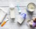 Baby feeding items on a wooden surface: two baby bottles filled with milk, a can of baby formula with a blue scoop, a small white towel, a white spoon with an orange handle, and a plush toy dog wearing a purple vest.