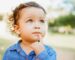 A young child with curly brown hair and light-colored eyes stands outdoors, wearing a denim shirt. The child looks thoughtful, as if pondering the benefits of choosing the right infant formula, with a finger resting on their chin, set against a blurred green background.